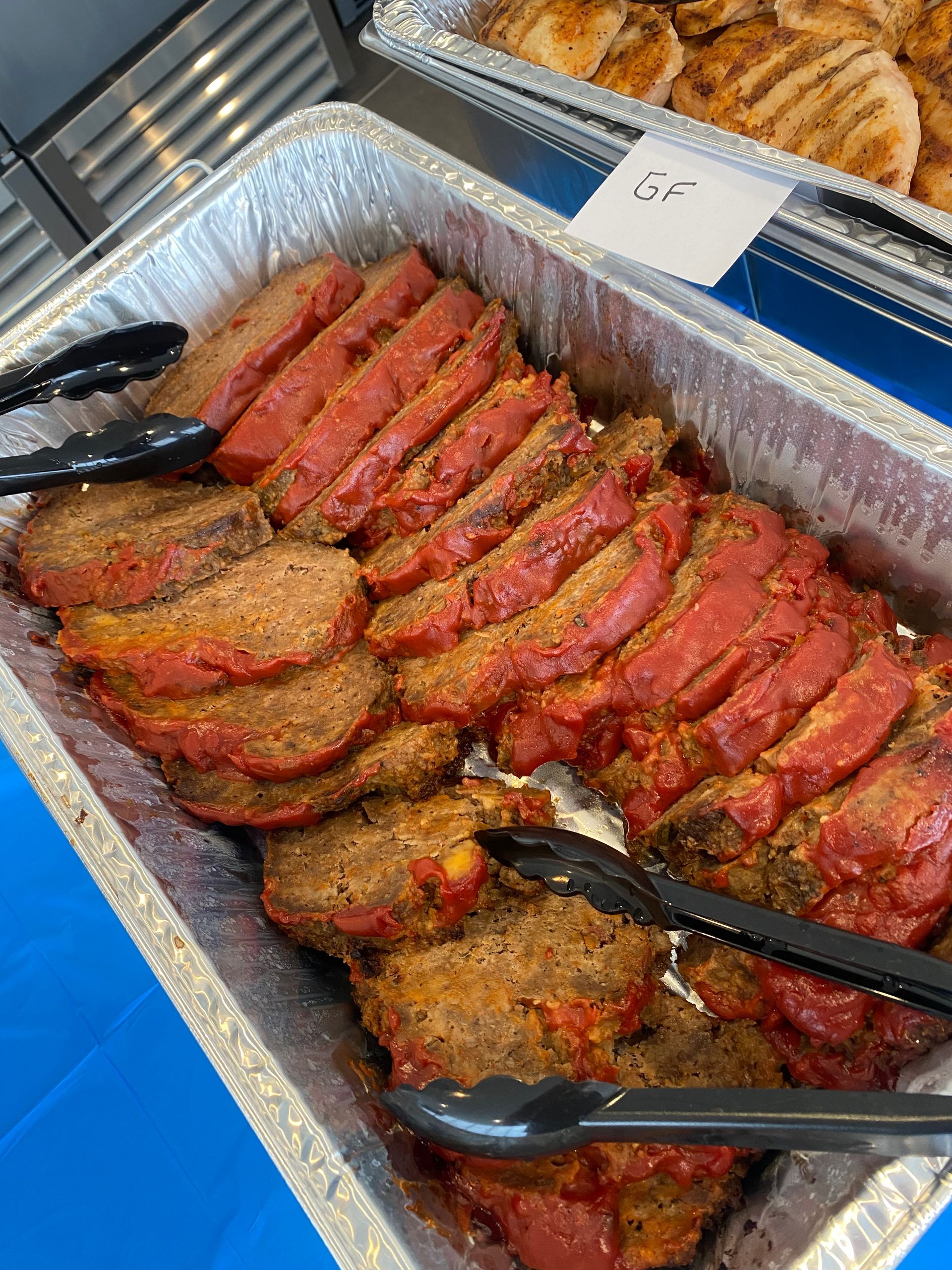 Sliced, red-seasoned meat in a foil tray with tongs; another tray of chicken visible in background.
