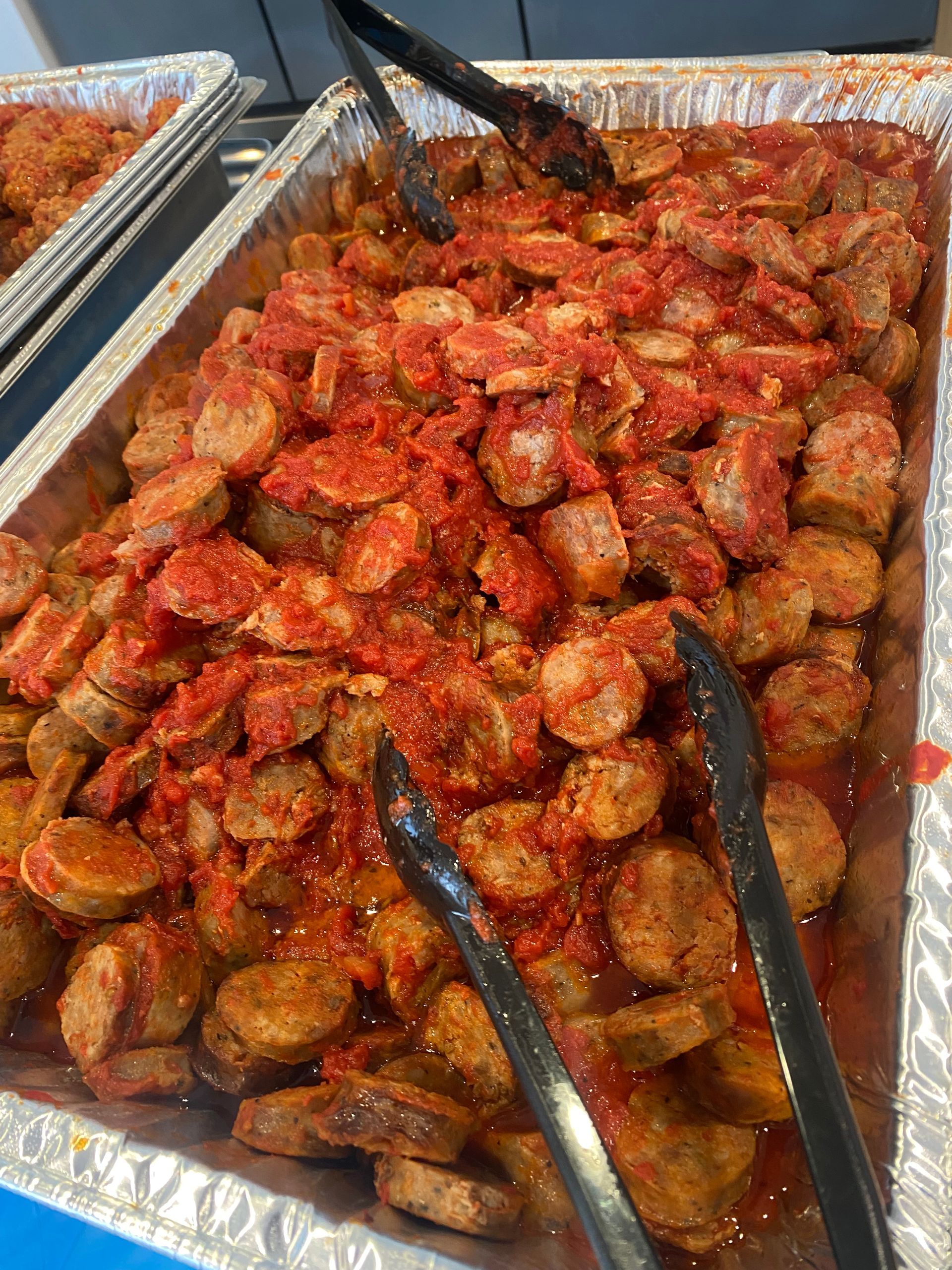 Sausage in tomato sauce in a foil pan, with serving tongs.