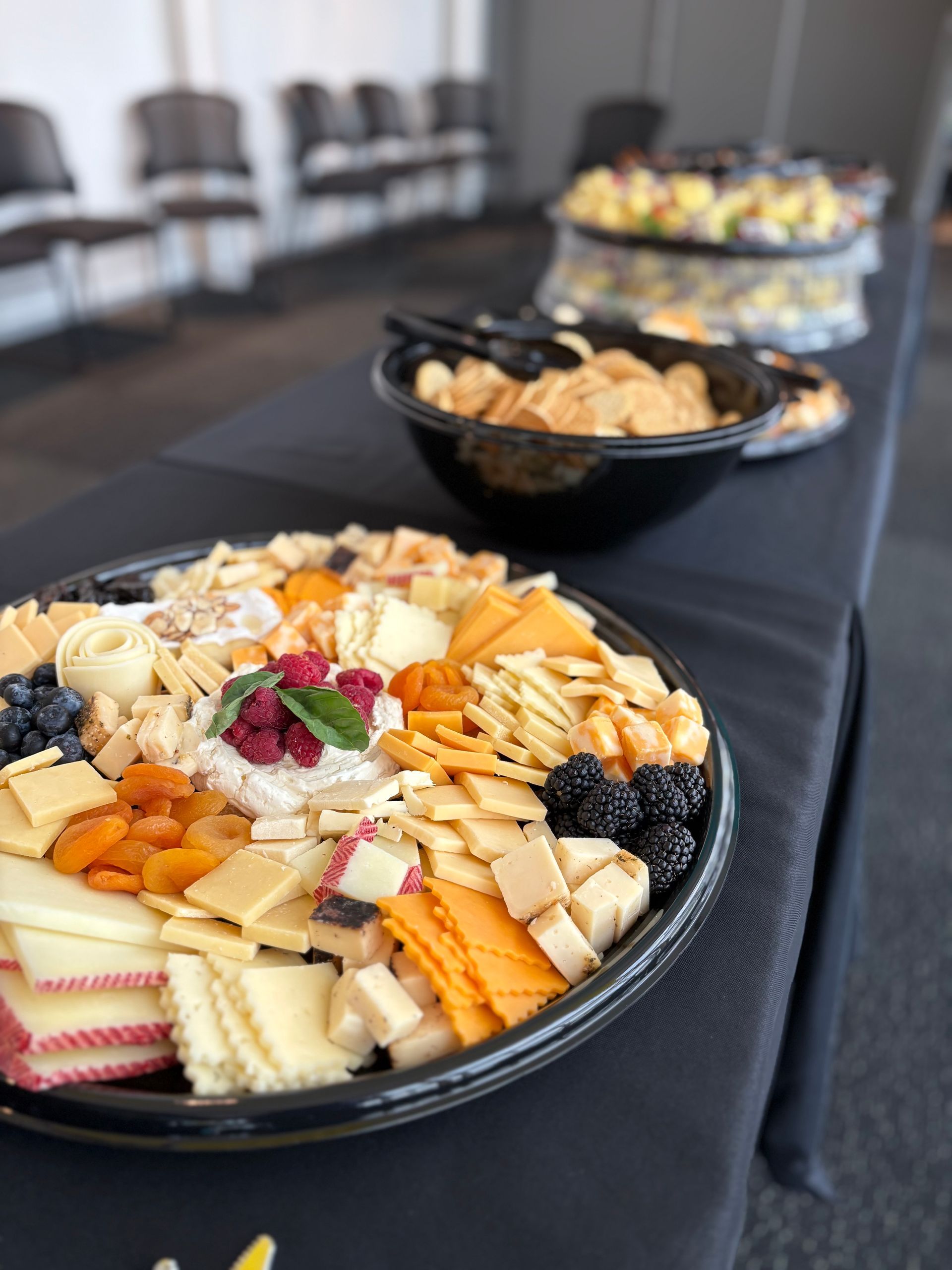 Charcuterie platter with cheeses, fruit, crackers on a black-covered table, with bowls of food in the background.
