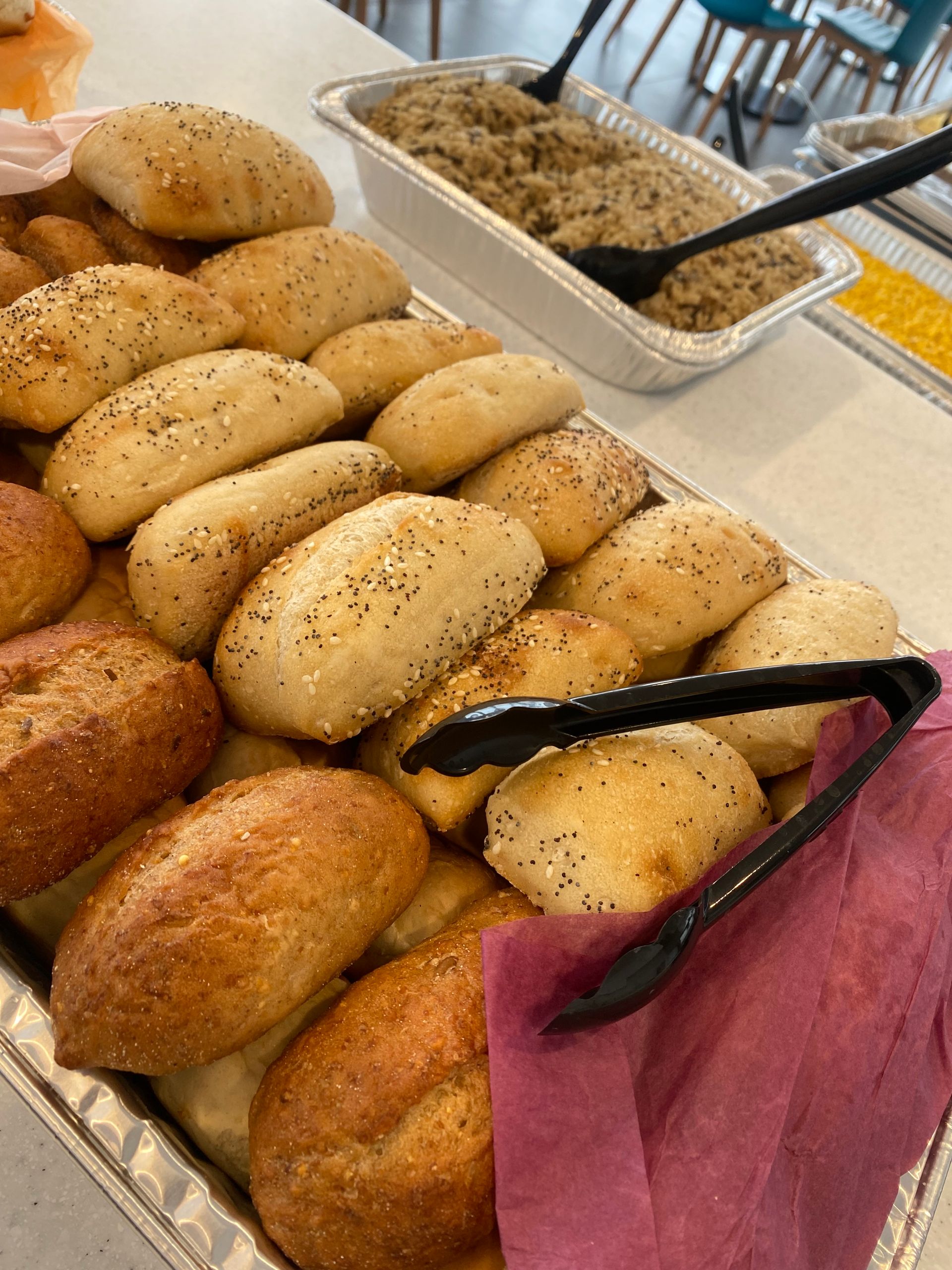 Tray of rolls, some with seeds, next to a dish of grain and tongs on a buffet.