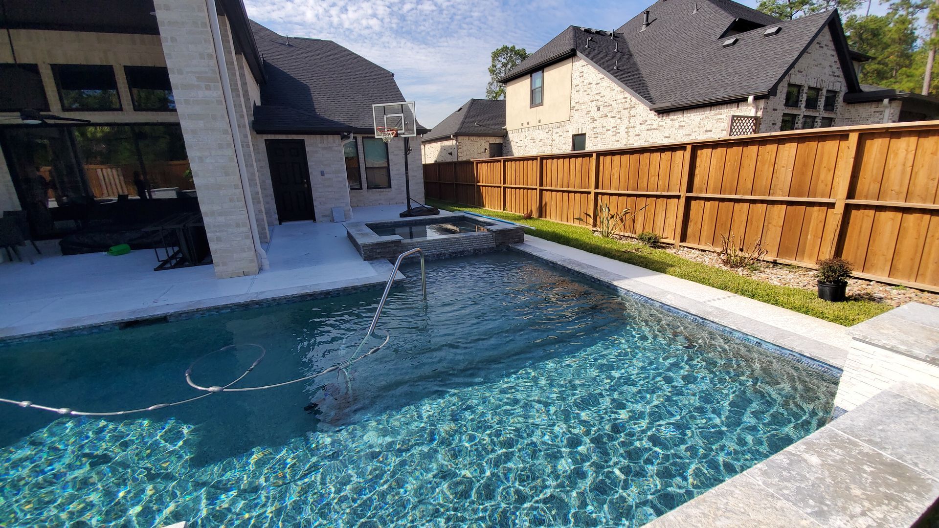 Backyard swimming pool with clear blue water, stone patio, waterfall feature, and wooden fence beside a house