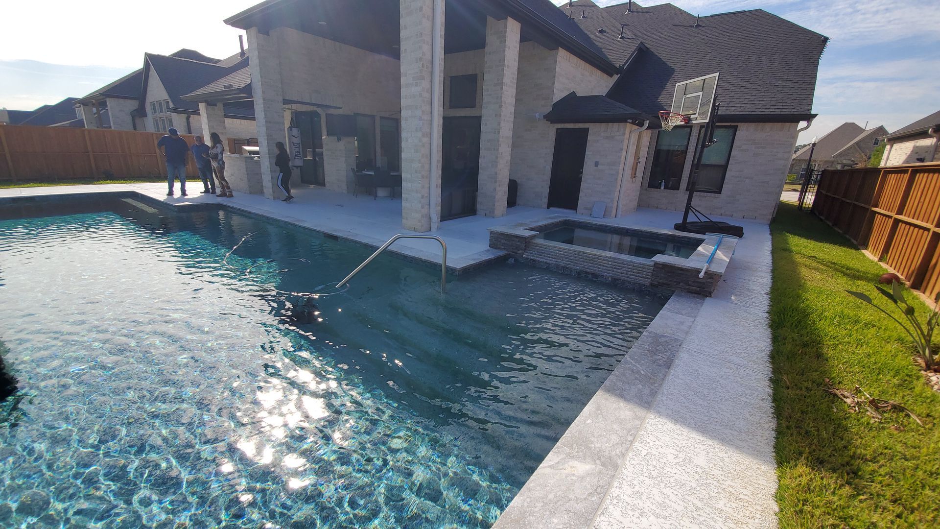Backyard swimming pool beside a covered patio and stone house, with a rock waterfall feature.