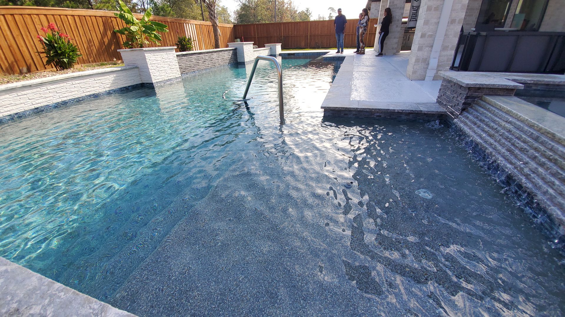 Backyard swimming pool with a waterfall, curved steps, and two people standing on the deck.