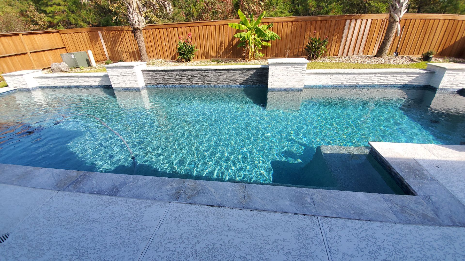 Backyard swimming pool with clear blue water, stone coping, and a wooden fence in the background