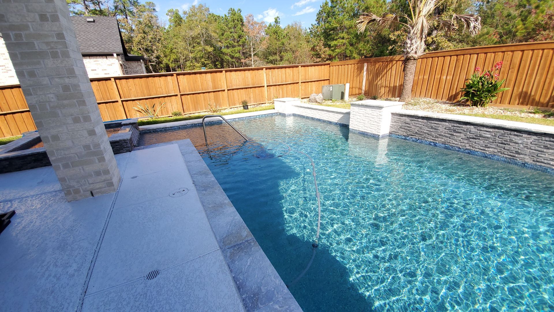Backyard swimming pool with blue water, stone patio, and wooden fence under sunny sky
