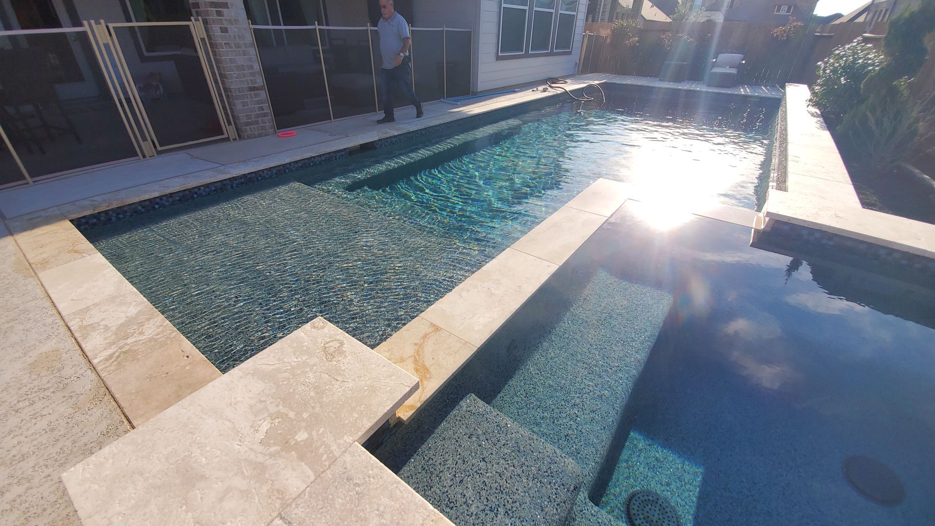 Sunlit backyard pool with clear water, stone deck, and a person standing near the far side.