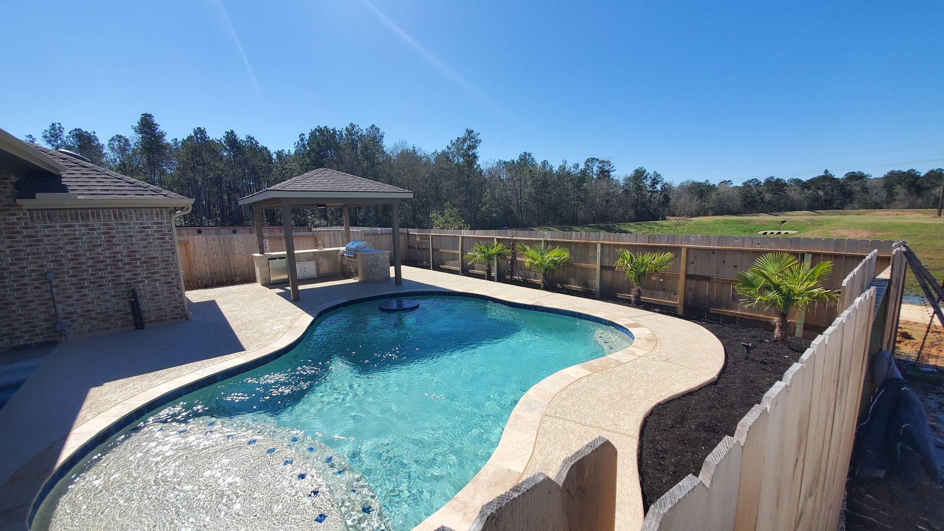 Backyard pool with clear blue water, stone patio, gazebo, and wooden fence under a sunny sky