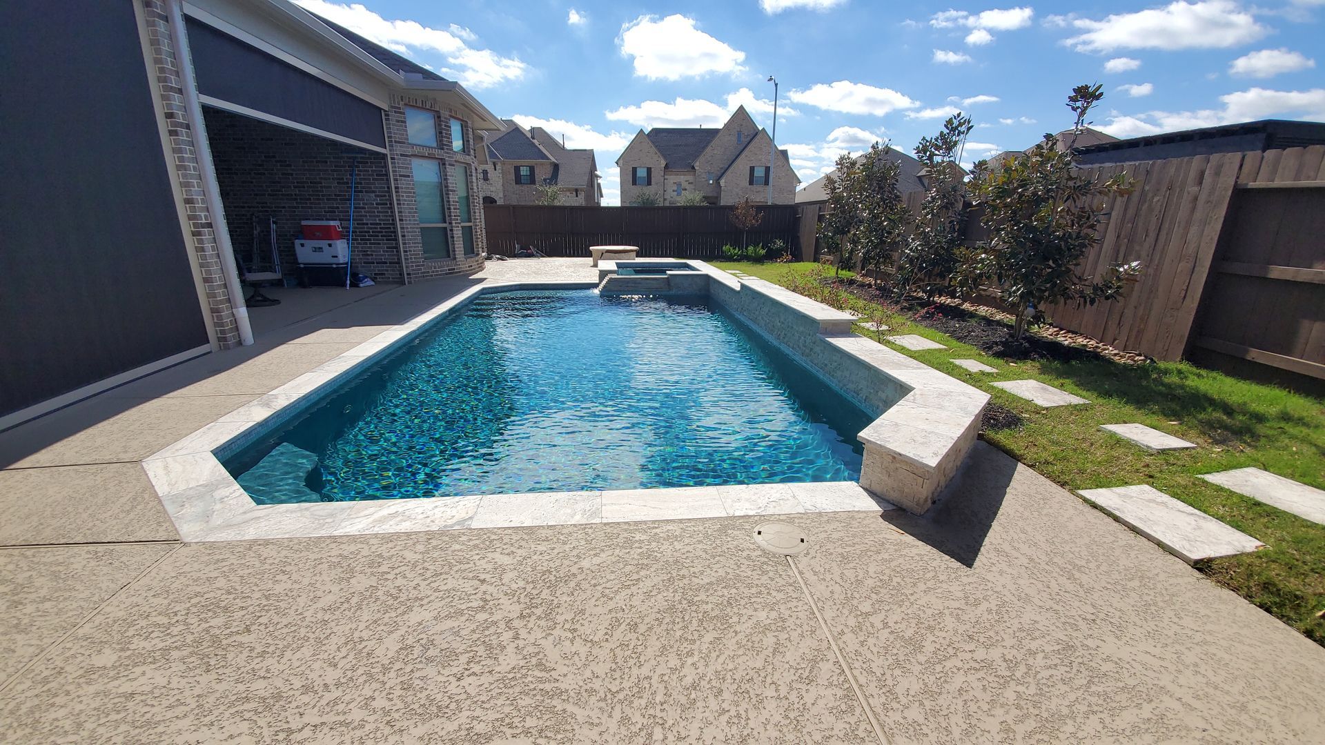 Backyard swimming pool with clear blue water, patio, lawn, and stone steps under a sunny sky