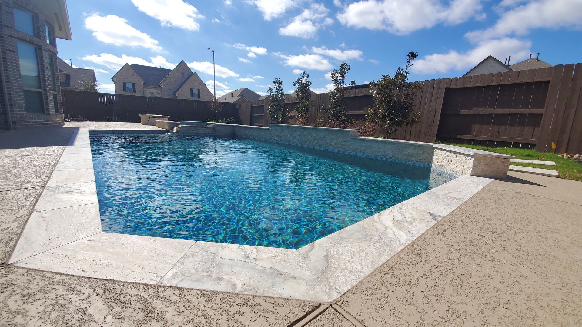 Backyard swimming pool with blue water, stone deck, and suburban houses under a partly cloudy sky
