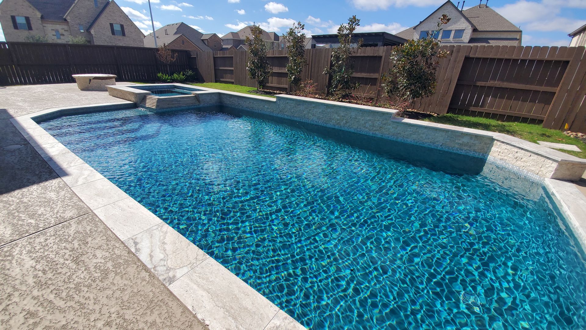 Backyard swimming pool with blue water, stone deck, and suburban houses in the background