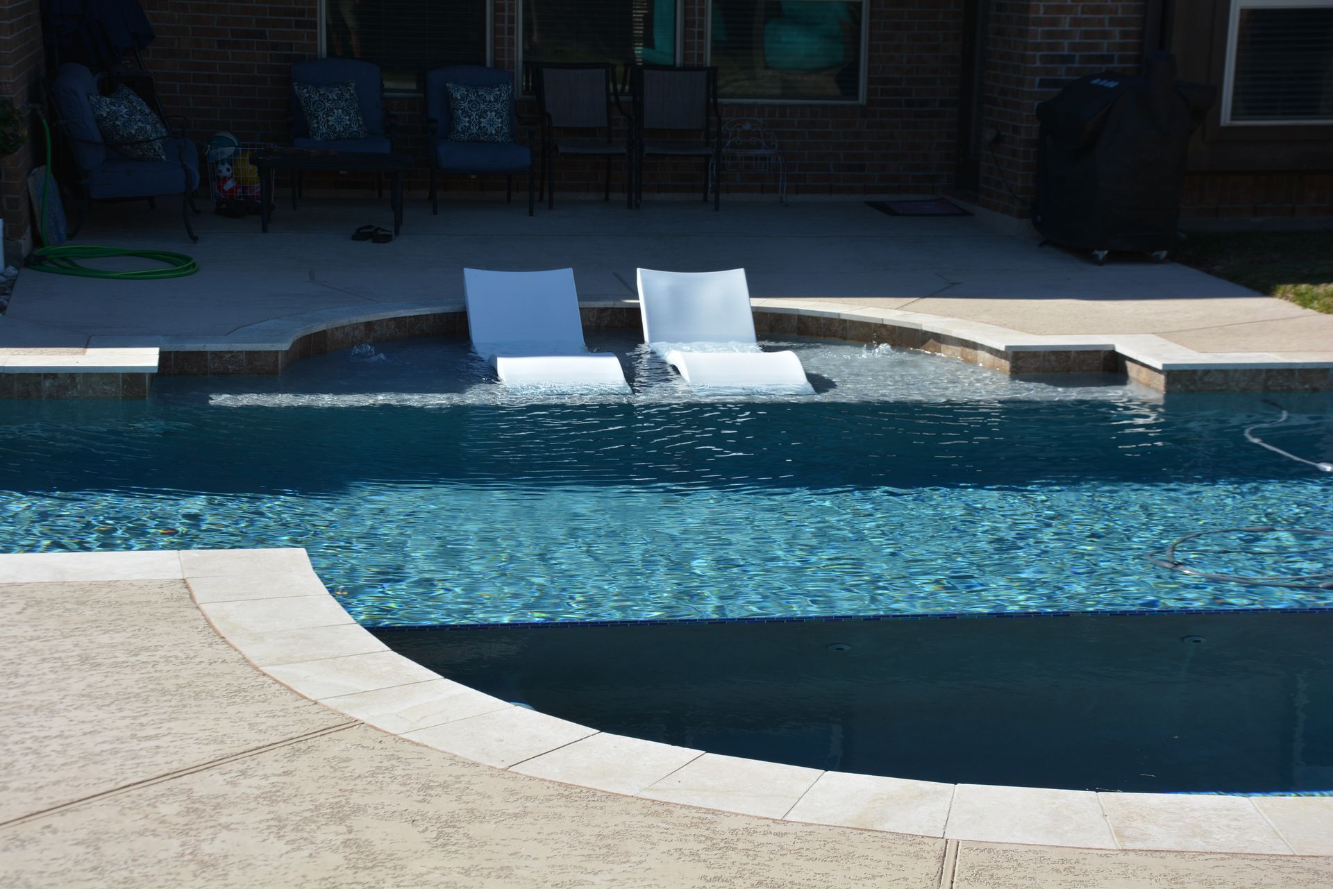 Backyard pool with two white lounge chairs on a stone deck at the water’s edge