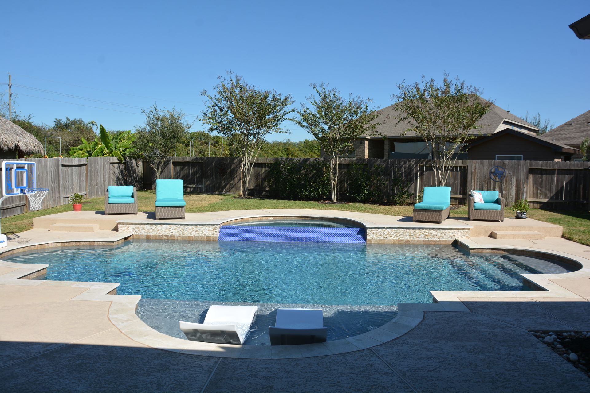 Backyard swimming pool with blue lounge chairs and a stone deck under a clear sky