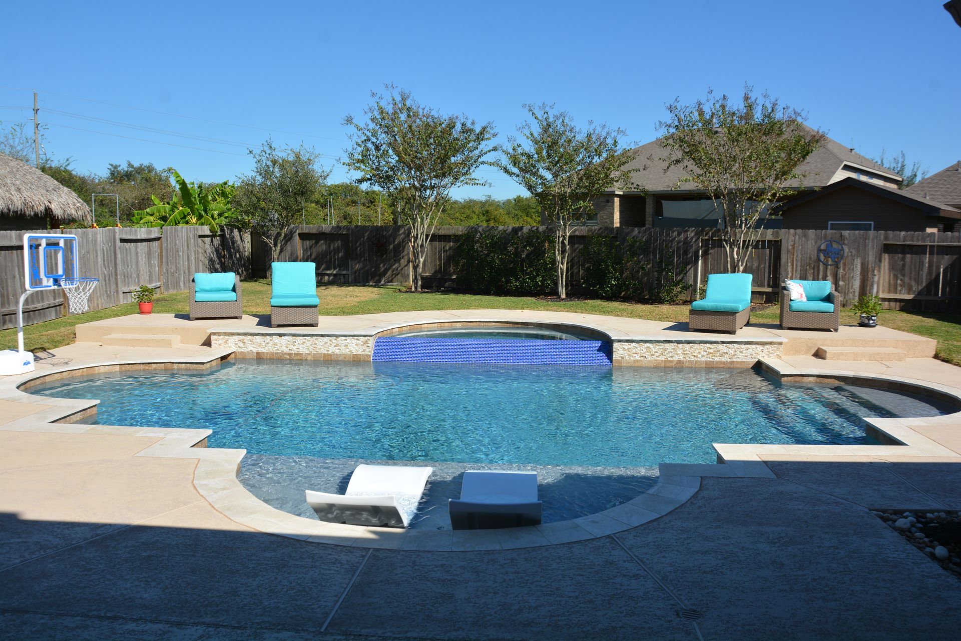 Backyard swimming pool with blue lounge chairs, stone patio, and surrounding trees and houses