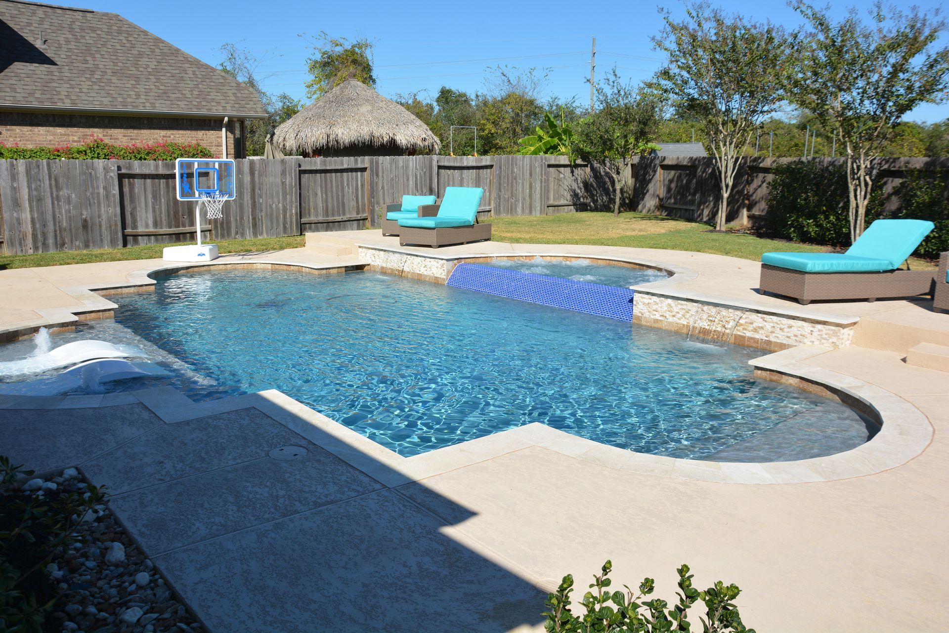 Backyard swimming pool with blue water, patio, lounge chairs, and a basketball hoop against a wooden fence