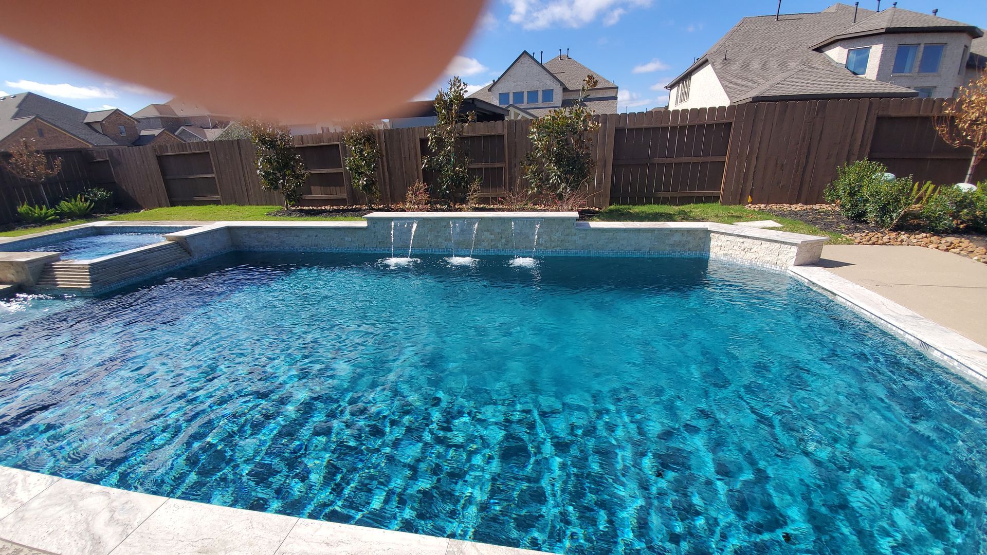 Backyard swimming pool with clear blue water, patio, fence, and houses in the background