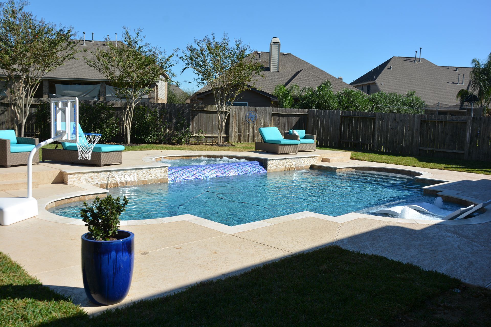 Backyard swimming pool with blue lounge chairs, potted plant, and fenced suburban homes in the background