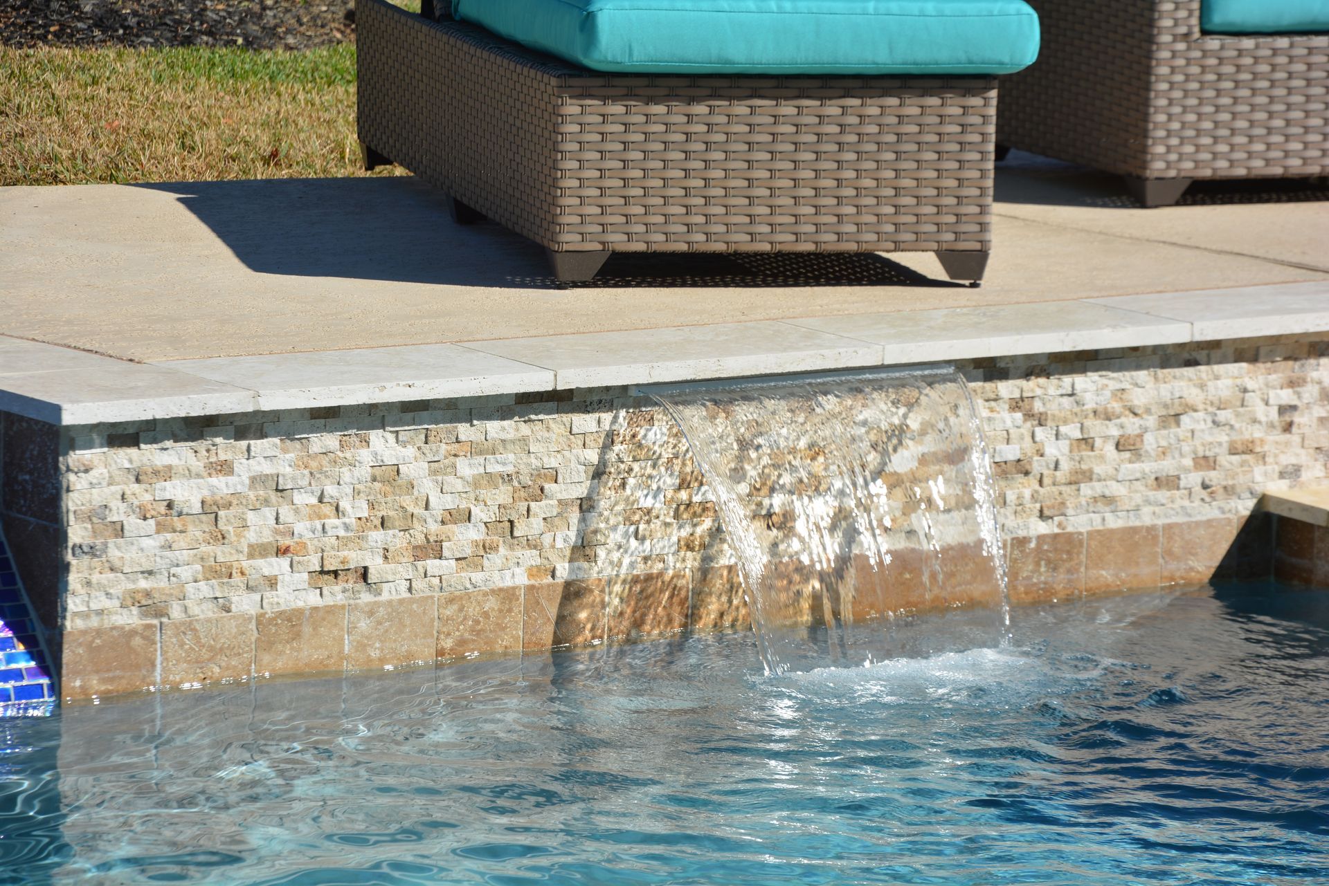 Poolside stone wall with water splashing into a blue swimming pool beside wicker lounge seating