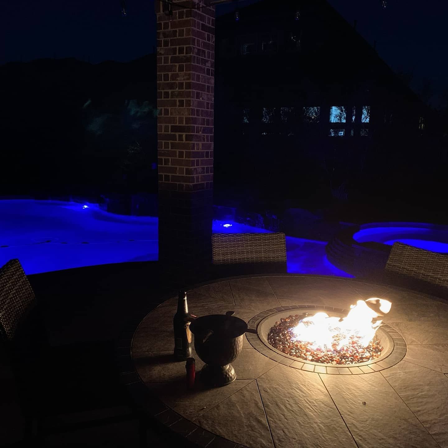 Night patio with a fire pit glowing beside a blue-lit pool and a brick pillar