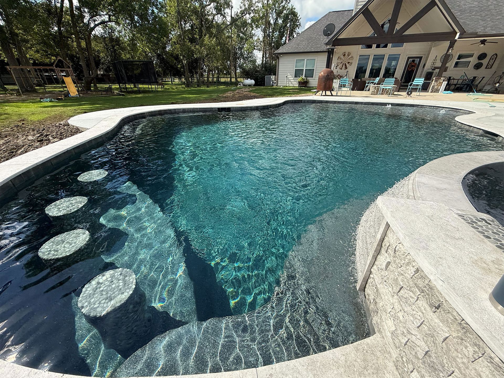 Backyard pool with turquoise water, stone edging, and a house with patio chairs in the background