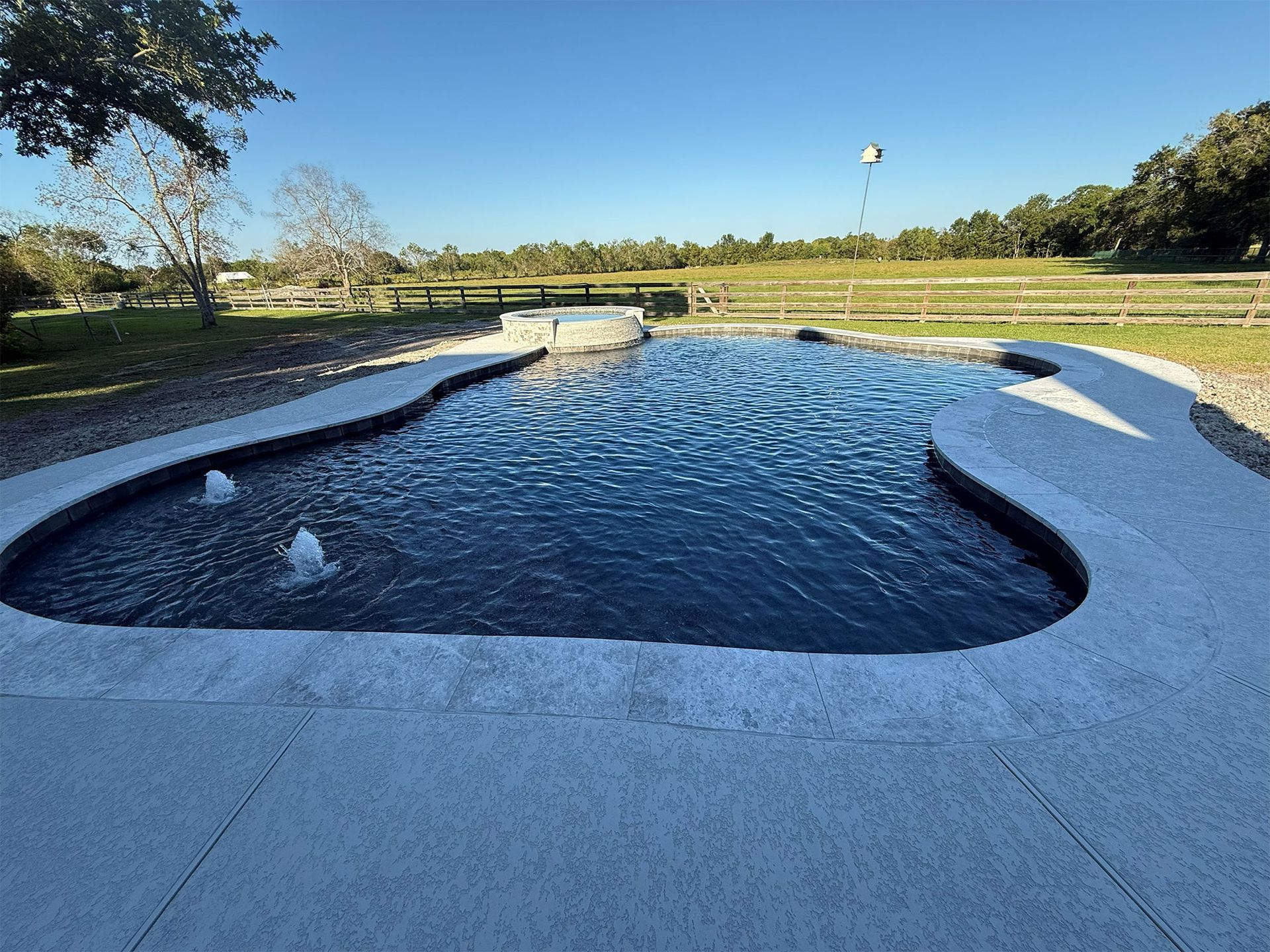 Curved backyard pool with dark water, white deck, and a grassy landscape under a clear blue sky