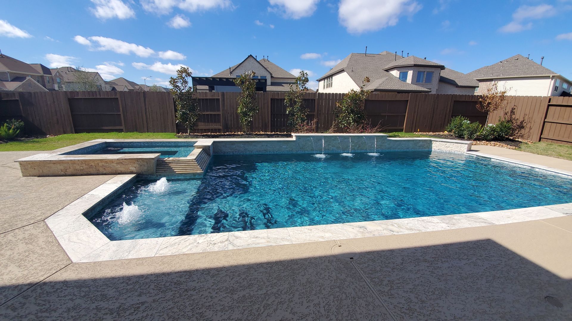 Backyard swimming pool with spa, white deck, and desert homes under a blue sky.