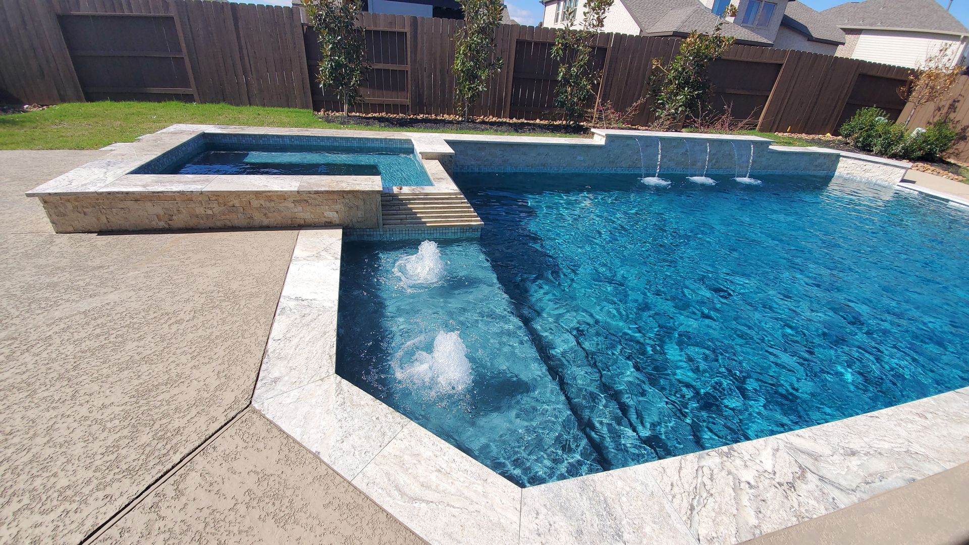 Backyard pool with blue water, attached hot tub, and beige stone deck under a sunny sky