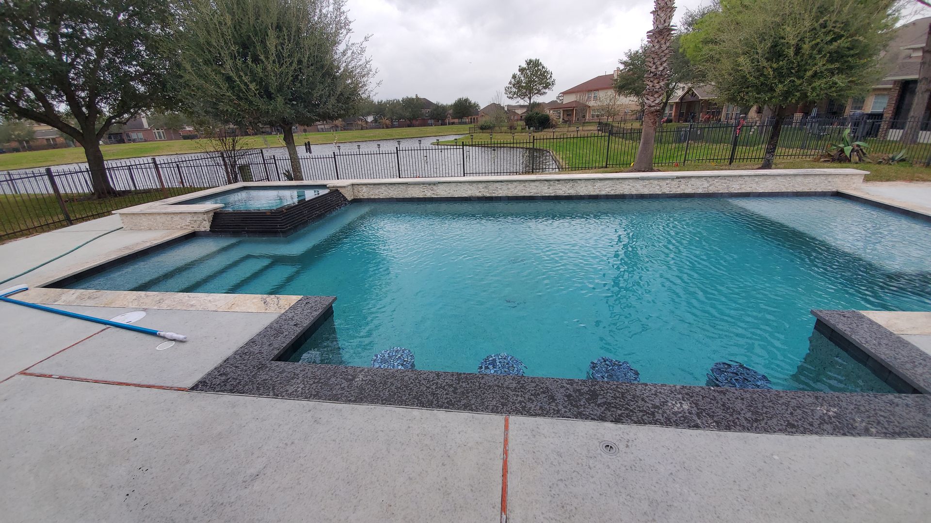 Empty backyard swimming pool with turquoise water, stone deck, and fence under cloudy sky