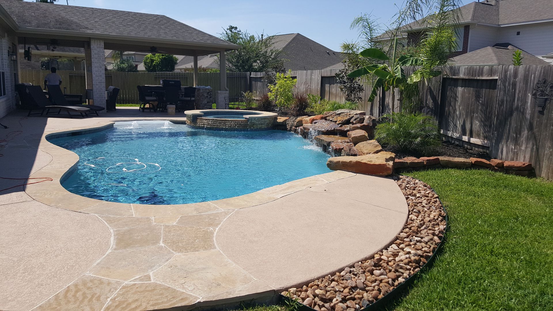 Backyard pool with stone patio, waterfall rocks, and green lawn under a covered patio
