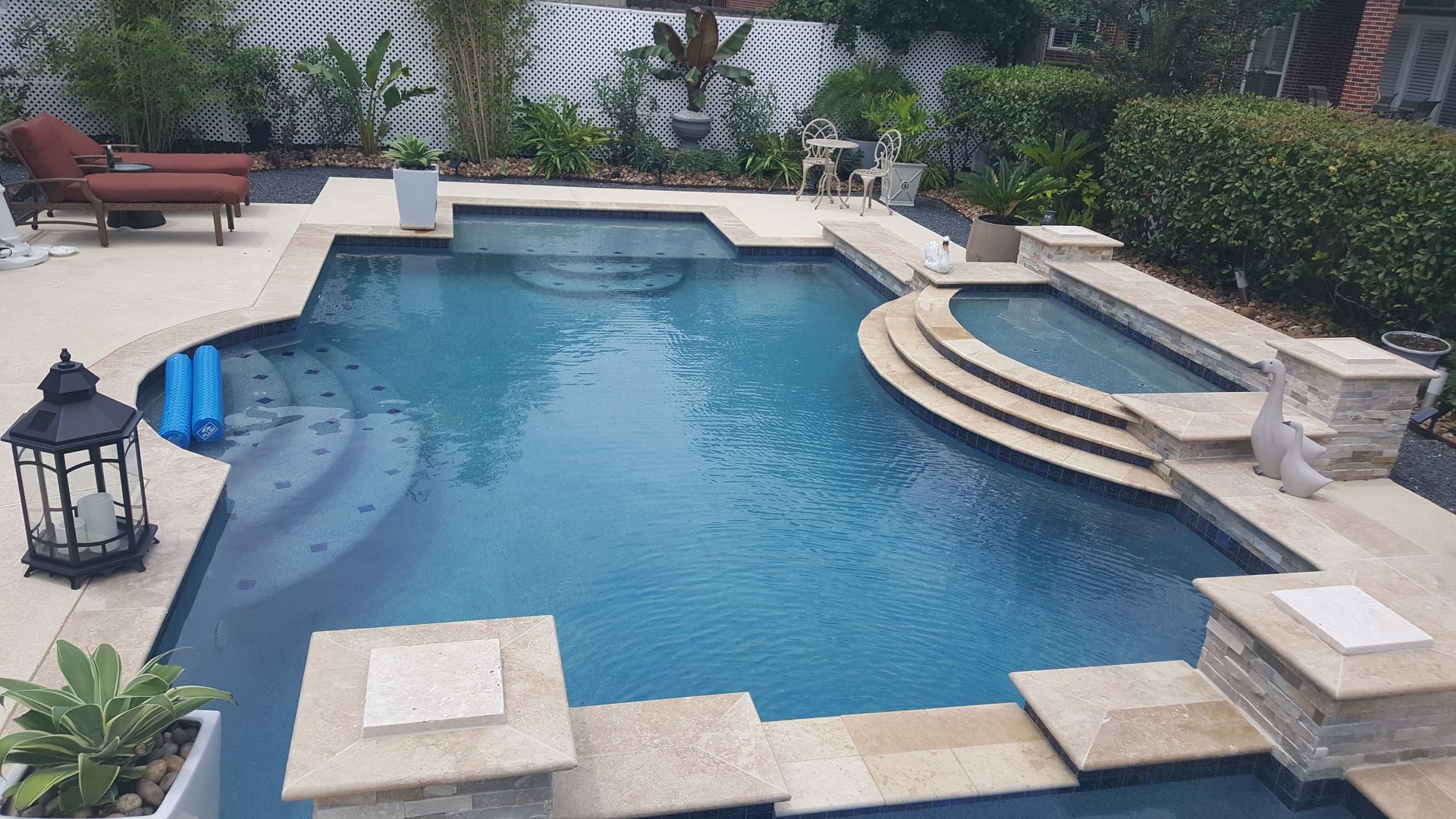 Backyard swimming pool with blue water, stone coping, lounge chairs, and potted plants around the deck