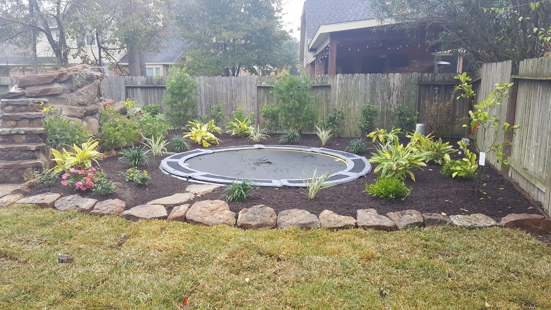 Backyard garden with a small circular pond, stone border, and colorful plants against a wooden fence