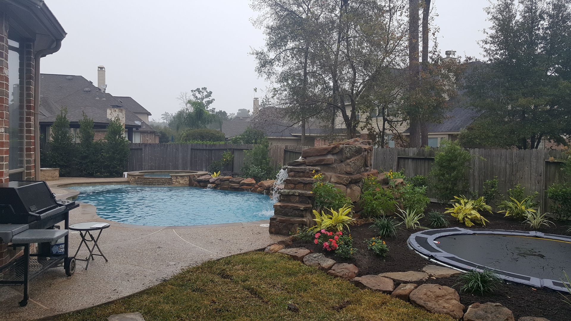 Backyard pool with patio chairs, trampoline, and flower beds on a cloudy day