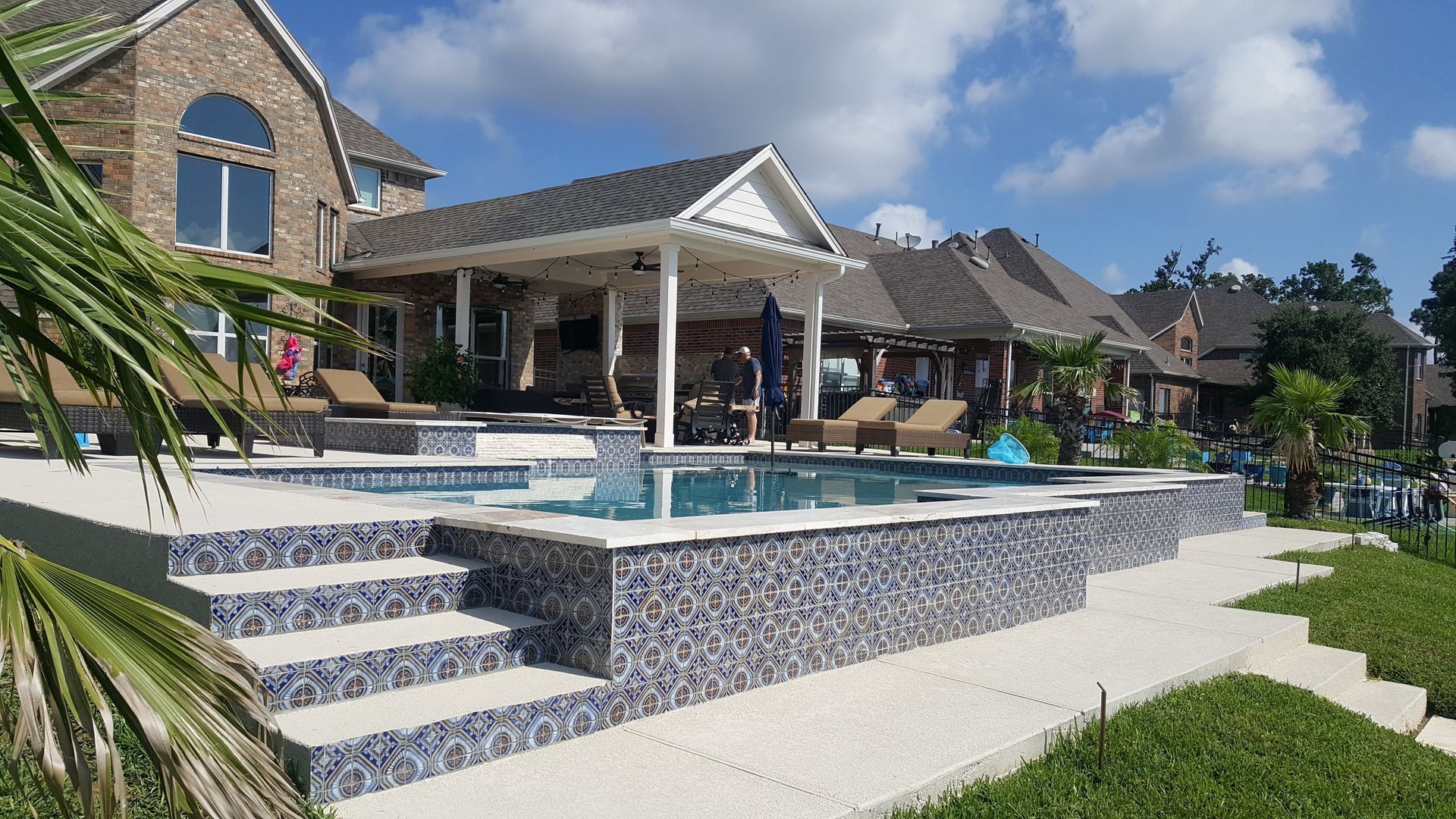 Luxury backyard pool with tiled steps, covered patio, and manicured lawn under a bright blue sky