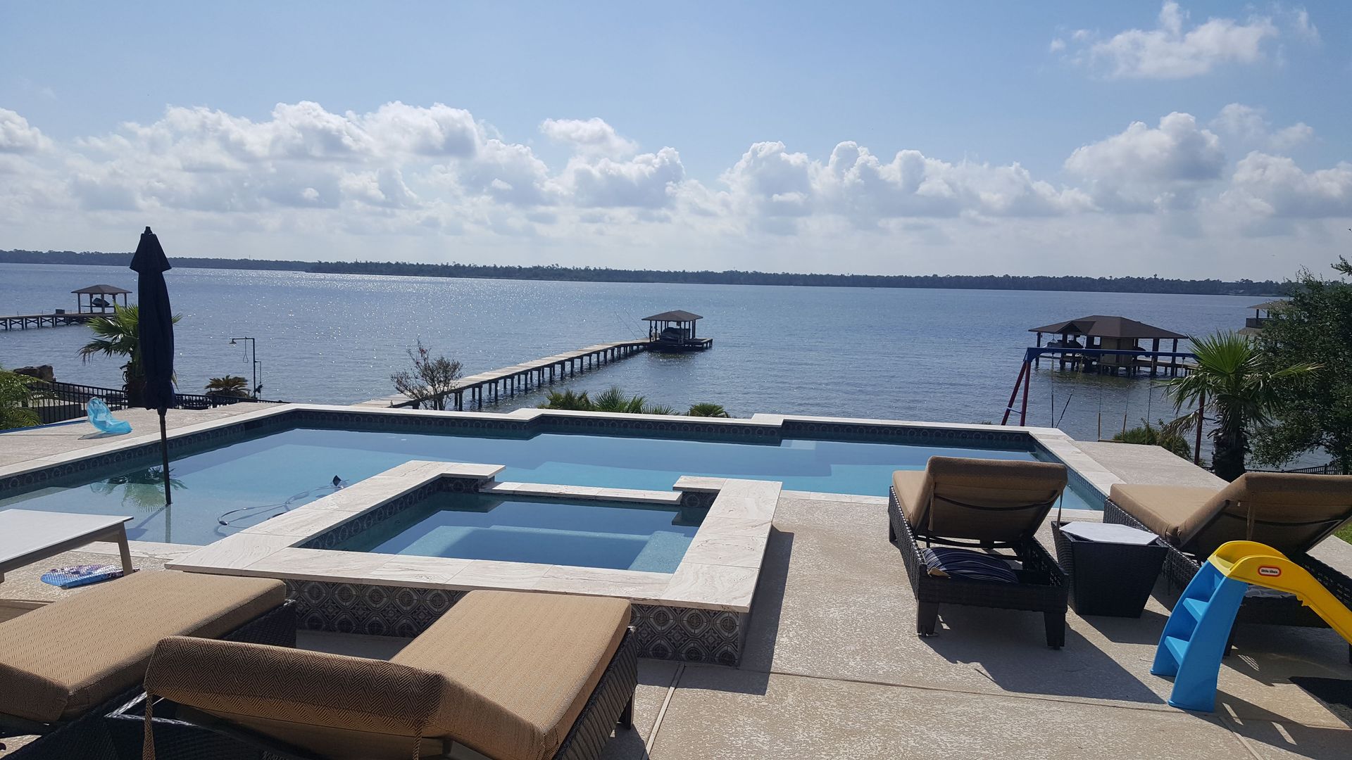 Infinity pool overlooking a blue bay, with lounge chairs and a pier under a partly cloudy sky