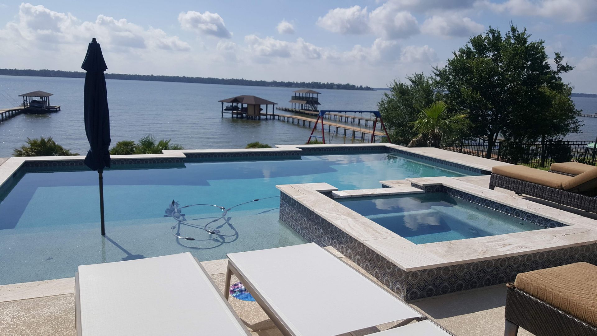 Infinity pool and hot tub overlooking a calm lake with a dock and partly cloudy sky