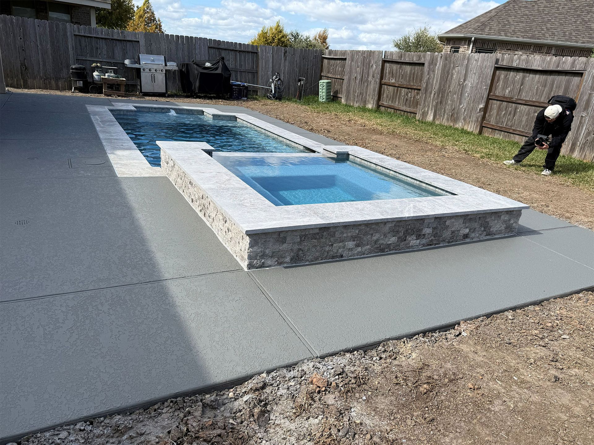 Rectangular backyard swimming pool with gray deck and a person standing near the fence