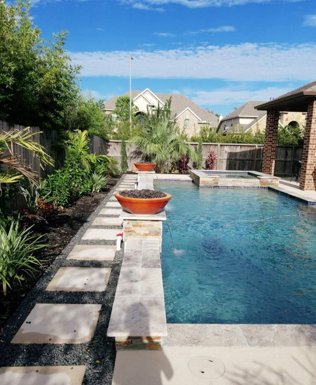 Backyard pool with stone walkway, potted plants, and houses under a blue sky