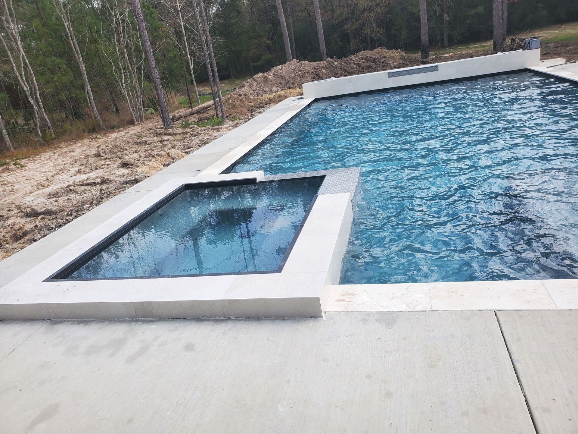 Modern backyard pool with a shallow tanning ledge and attached hot tub, bordered by white stone decking.
