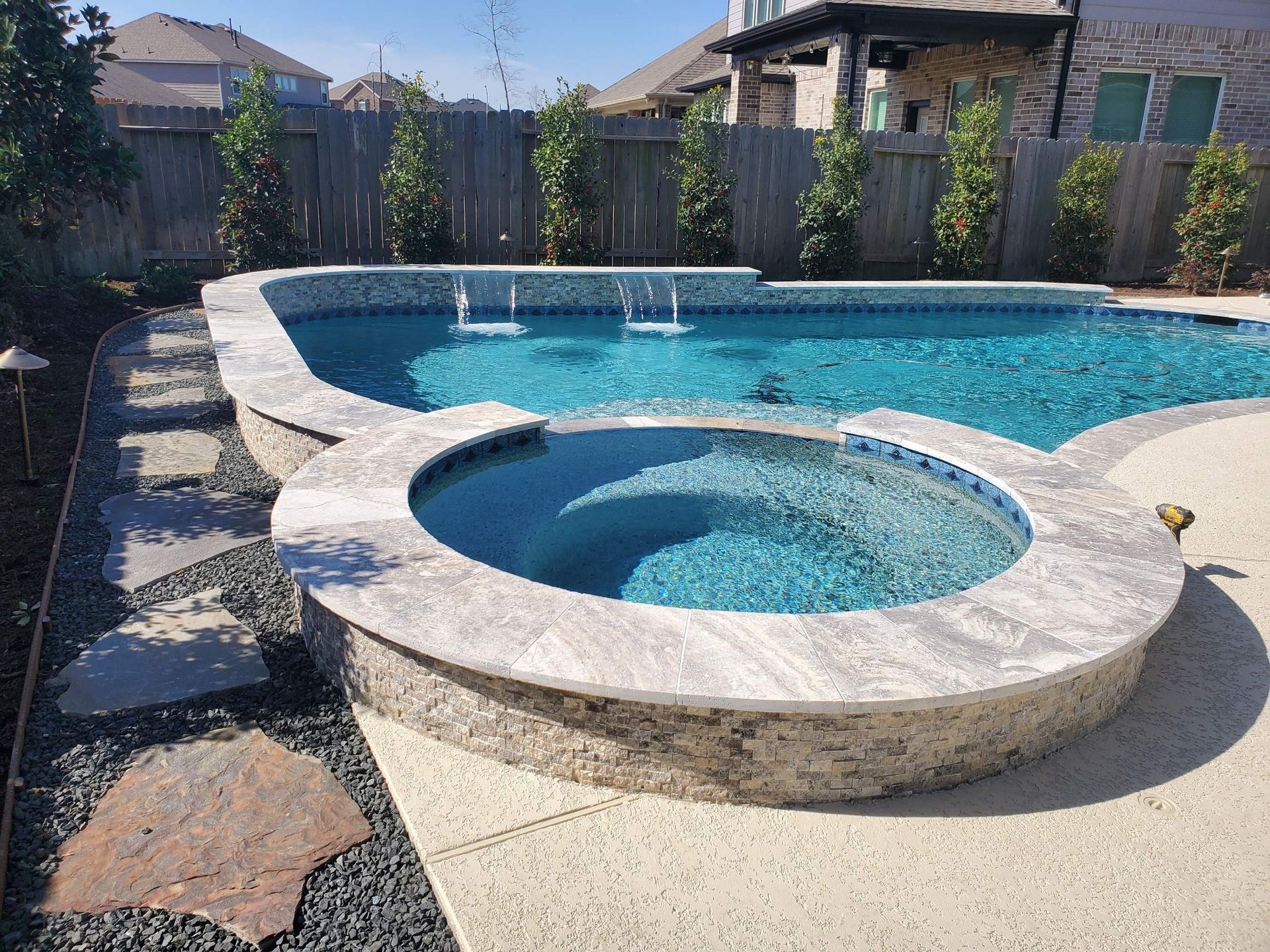 Backyard pool with attached hot tub, stone deck, and privacy fence under a clear sky