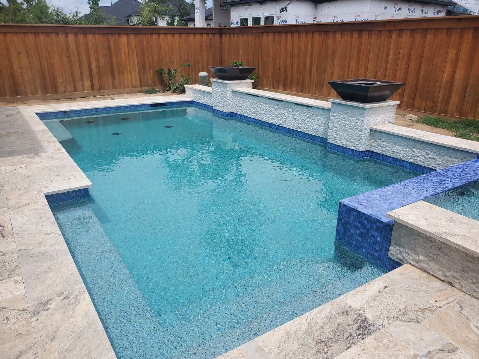 Modern backyard swimming pool with blue water, stone deck, and wooden privacy fence
