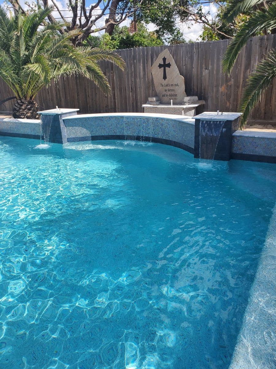 Blue backyard pool with stone waterfall and palm trees behind a privacy fence