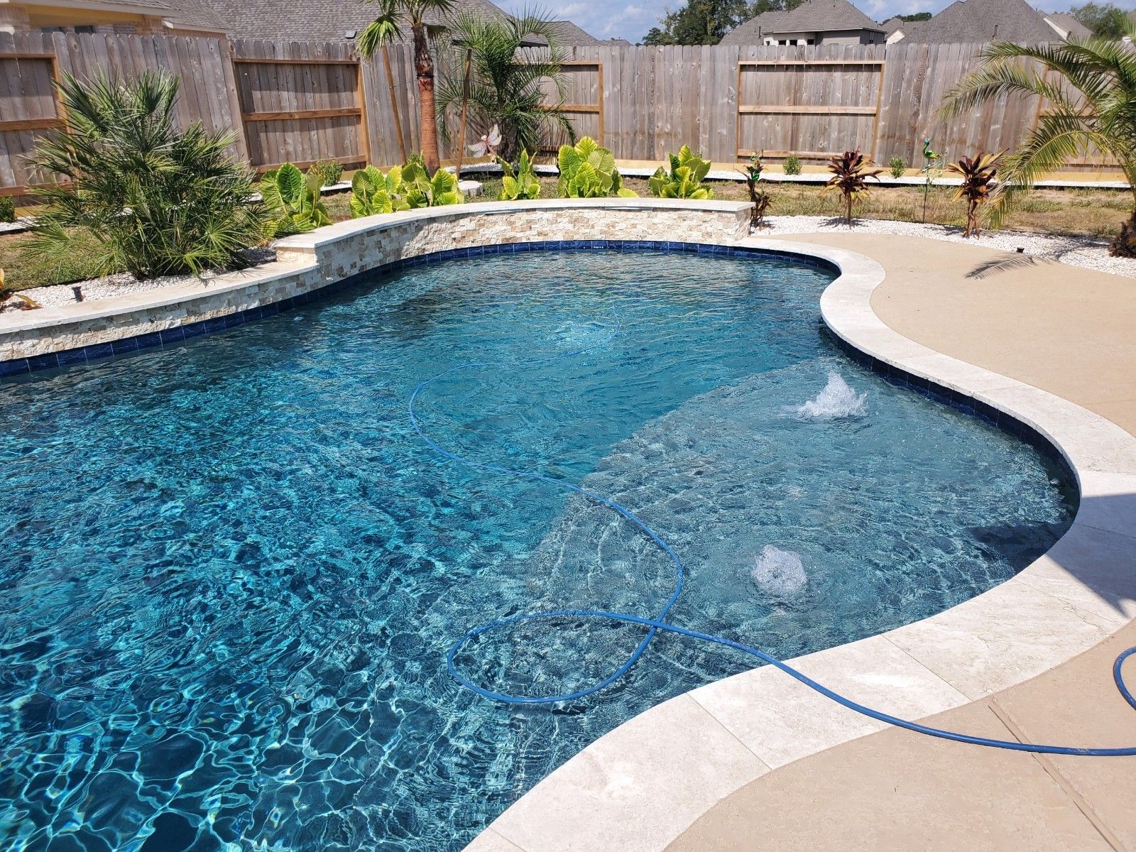 Curved backyard swimming pool with blue water, stone coping, beige deck, and wooden fence surrounding it