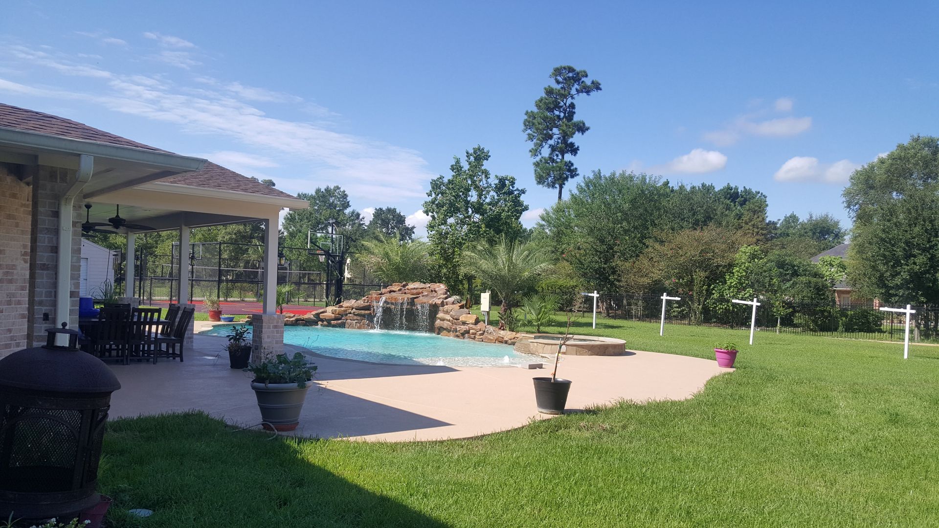 Backyard patio with a swimming pool, green lawn, potted plants, and trees under a blue sky.
