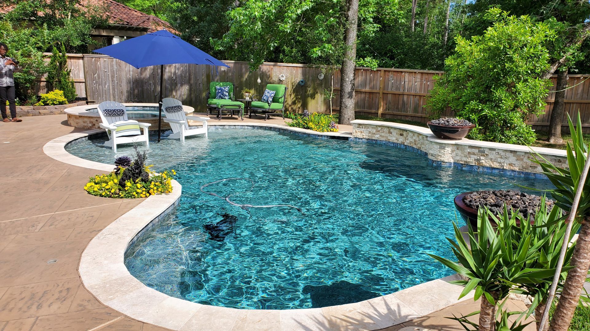 Backyard swimming pool with blue water, patio seating, and a blue umbrella surrounded by greenery