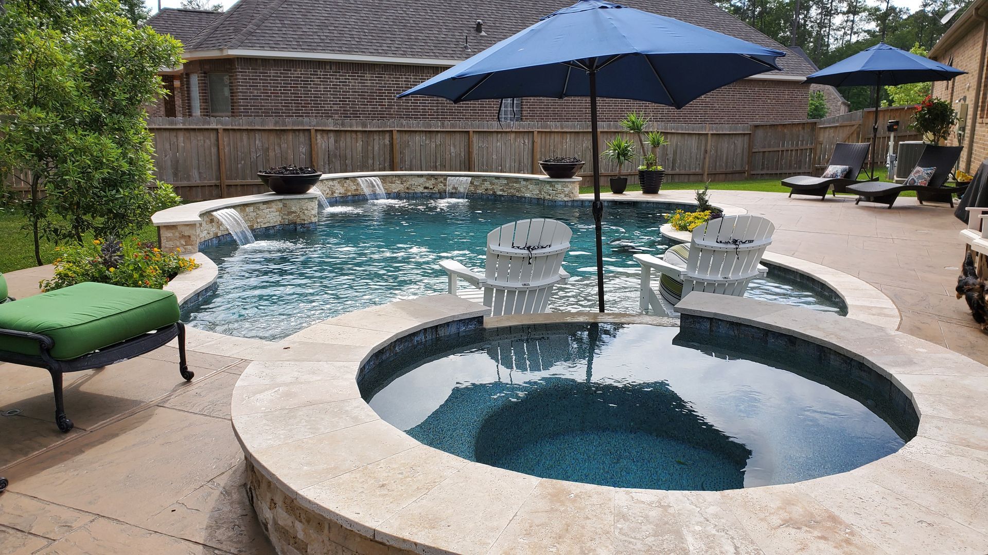 Backyard pool with a round hot tub, blue umbrellas, lounge chairs, and a stone patio