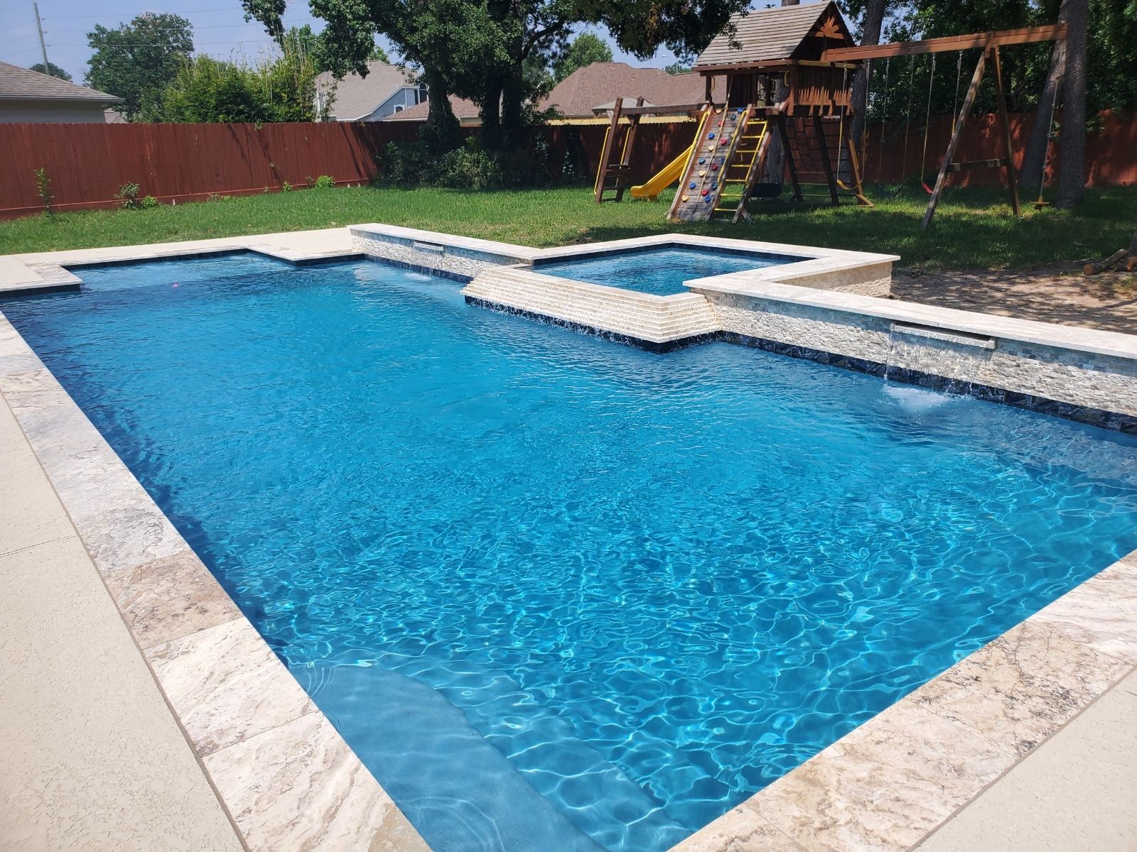 Backyard swimming pool with blue water, stone deck, and a play structure in the background