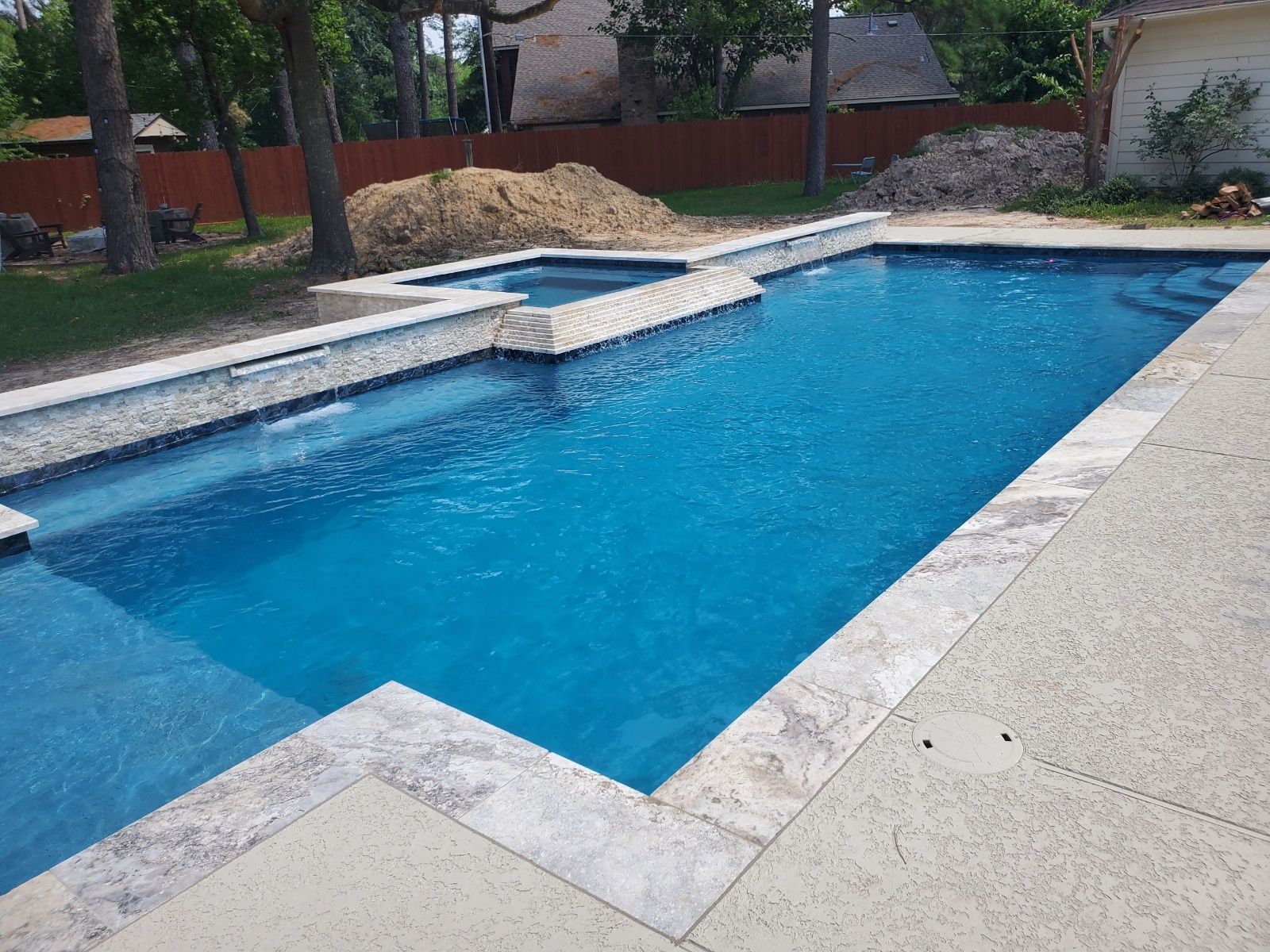 Backyard swimming pool with blue water, stone deck, and steps, surrounded by trees and a brick fence
