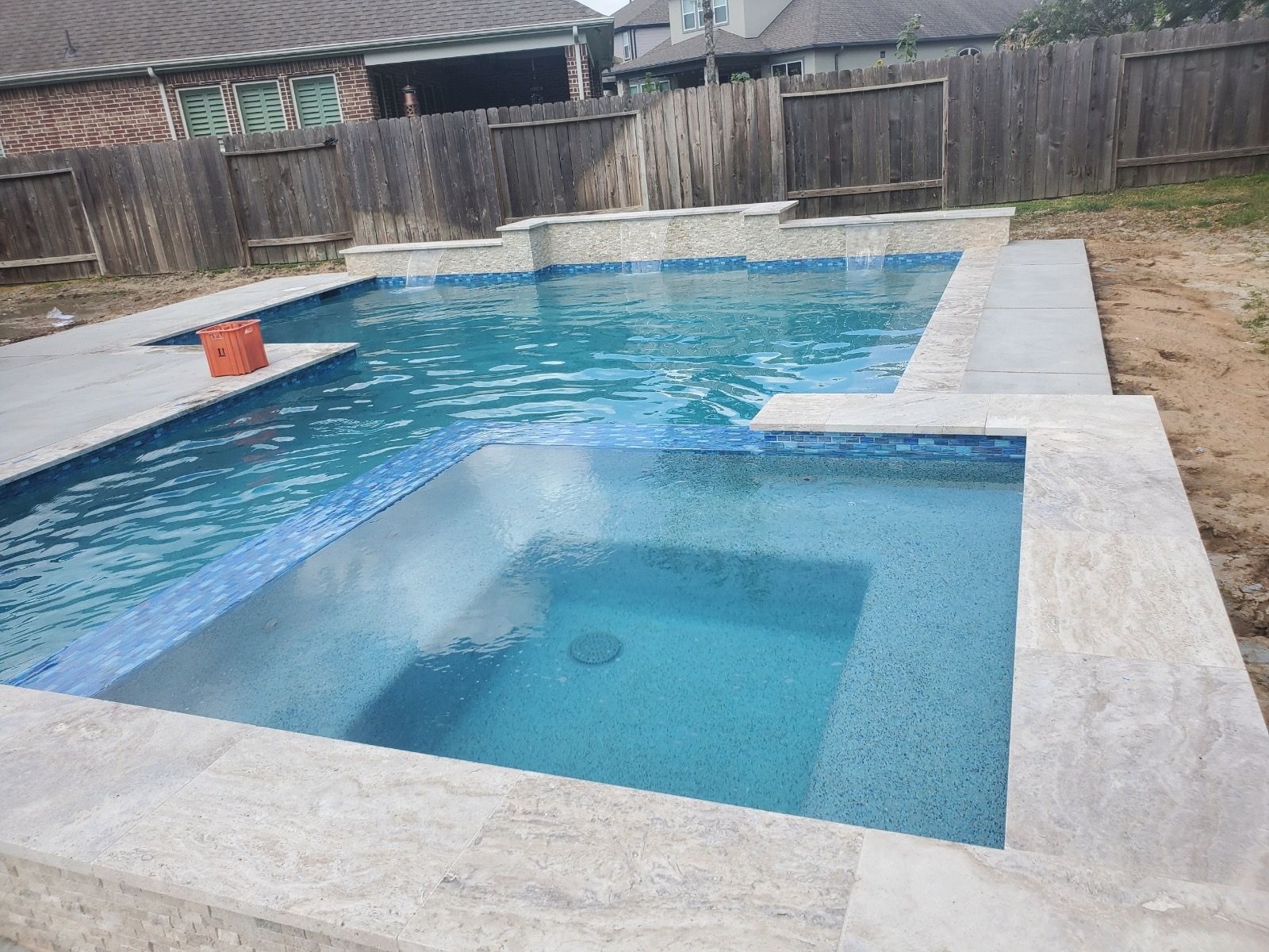 Backyard swimming pool with a raised square spa and blue water, surrounded by a concrete deck and wooden fence
