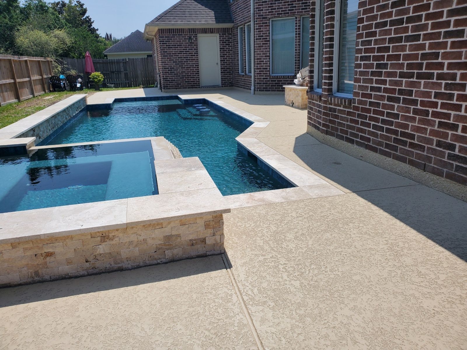 Backyard patio with a rectangular swimming pool, brick house, and stone deck in sunlight