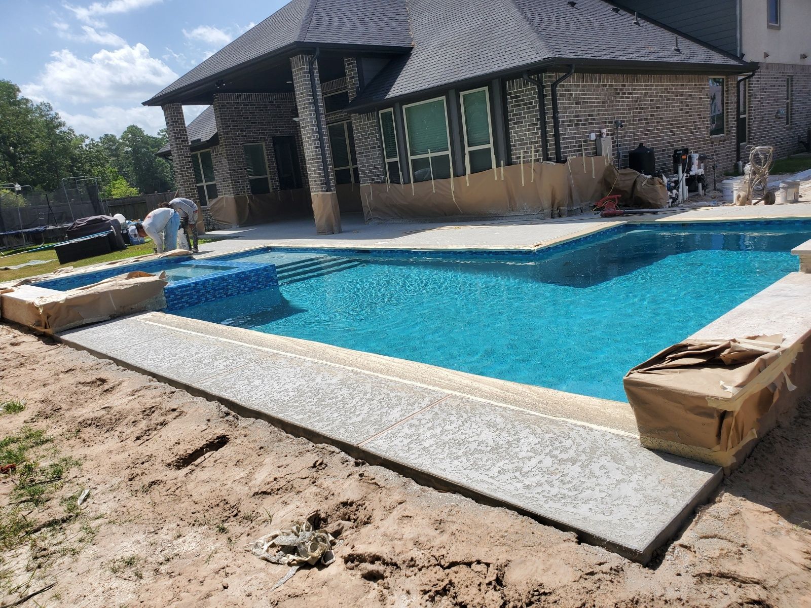 Backyard swimming pool beside a stone house, with workers and construction materials around it.