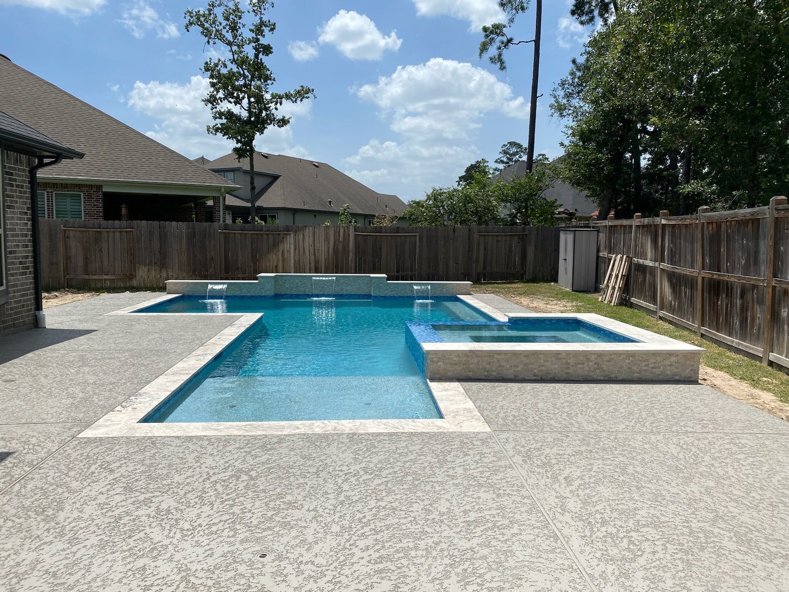 Backyard pool with blue water, concrete deck, wooden fence, and trees under a partly cloudy sky